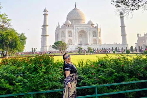 A woman posing in saree in front of the iconic Taj Mahal, surrounded by lush green gardens in Agra.